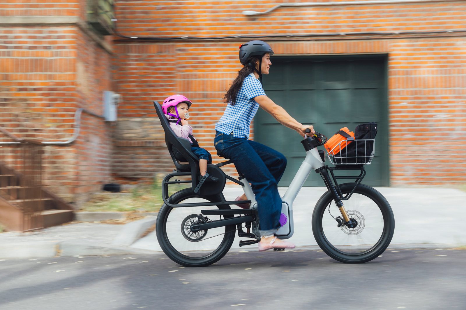 A rider carries a kid passenger on a TM-B past a brick building.