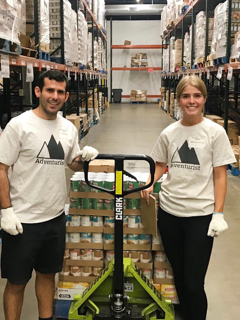 The two founders of Adventurist Backpack Co. in a Food Bank of the Rockies warehouse with a pallet of donated food.