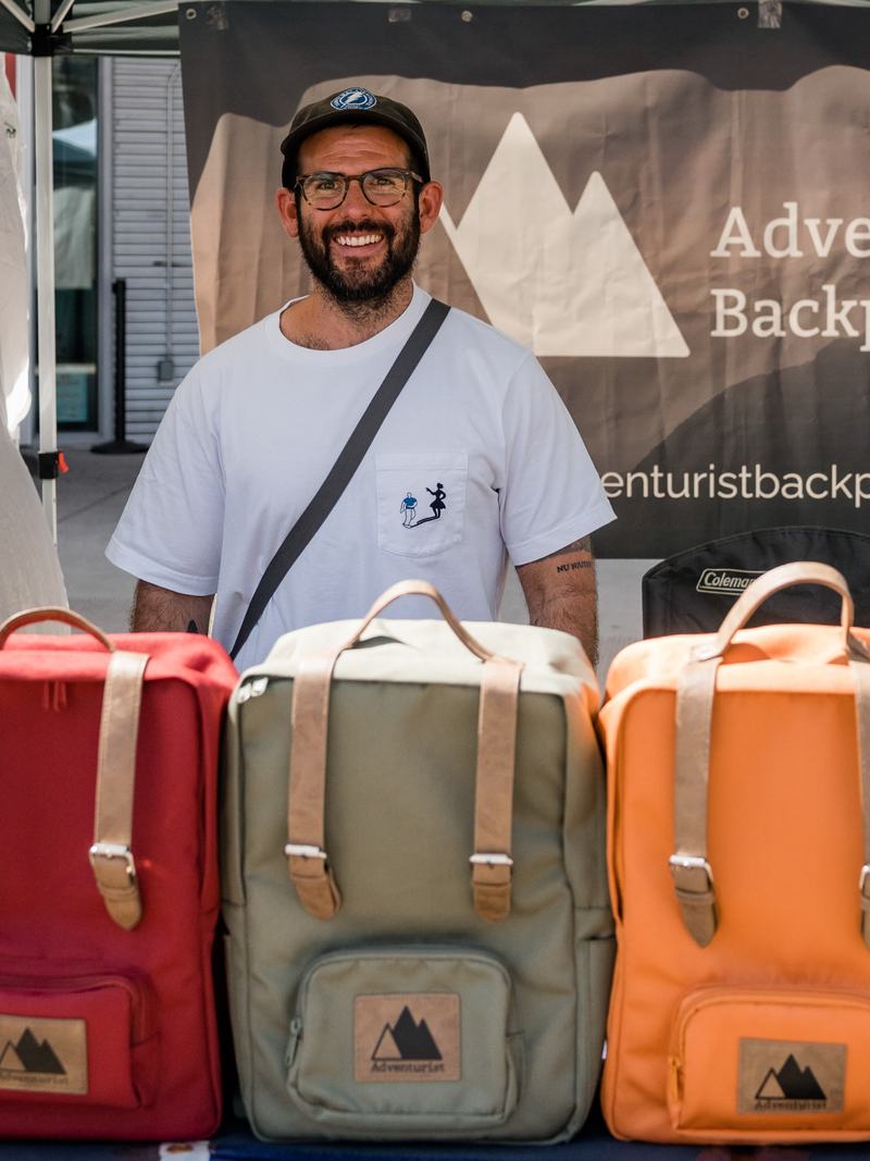 The founder of Adventurist Backpack Co. behind a row of five colorful backpacks at a market booth.