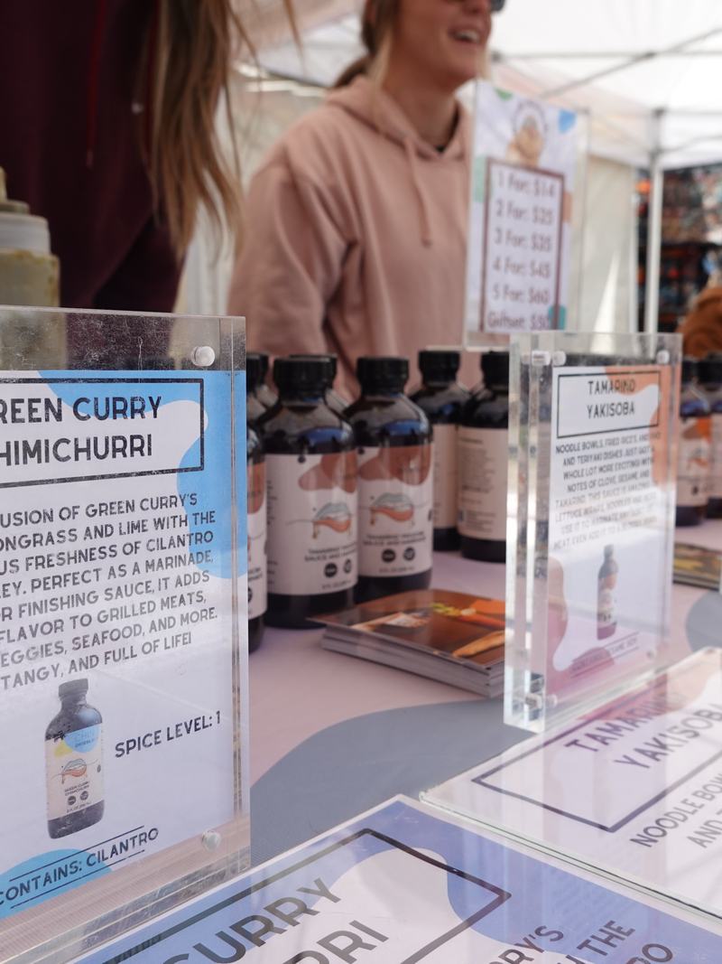 The Chin Dribblin' booth at a market, with sauce bottles and product cards including a Green Curry Chimichurri.