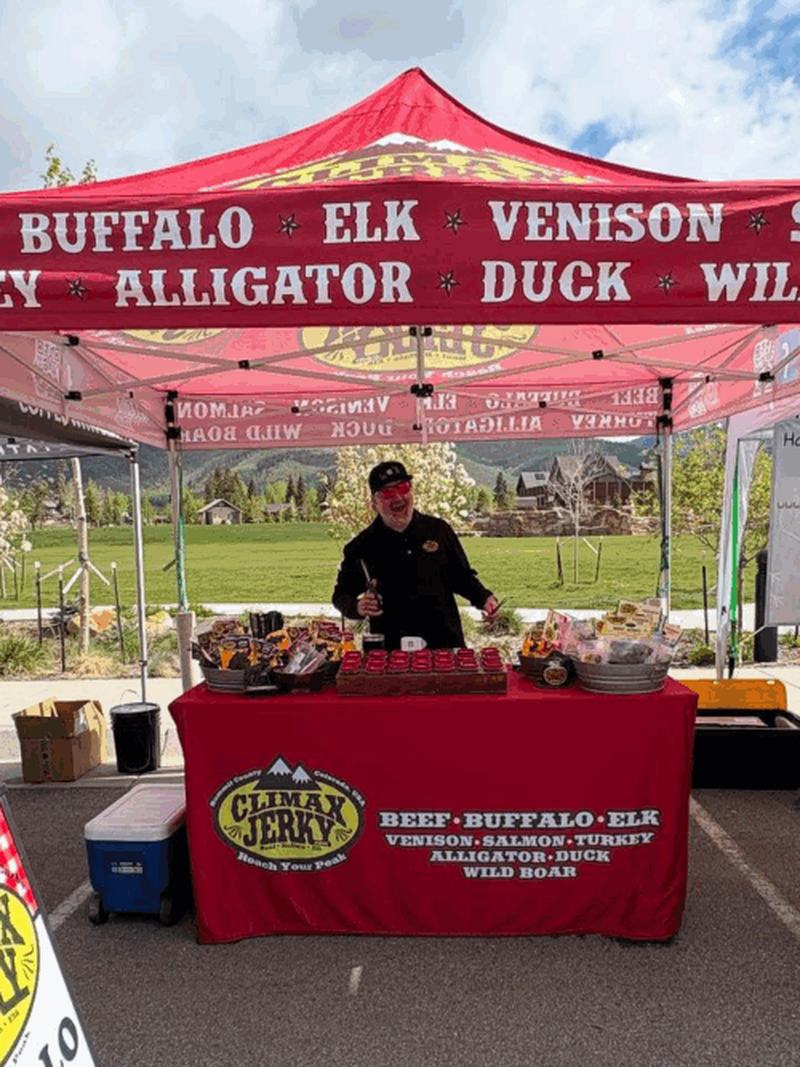 The Climax Jerky tent at a market, a banner listing buffalo, elk, and beef jerky above a table of product.