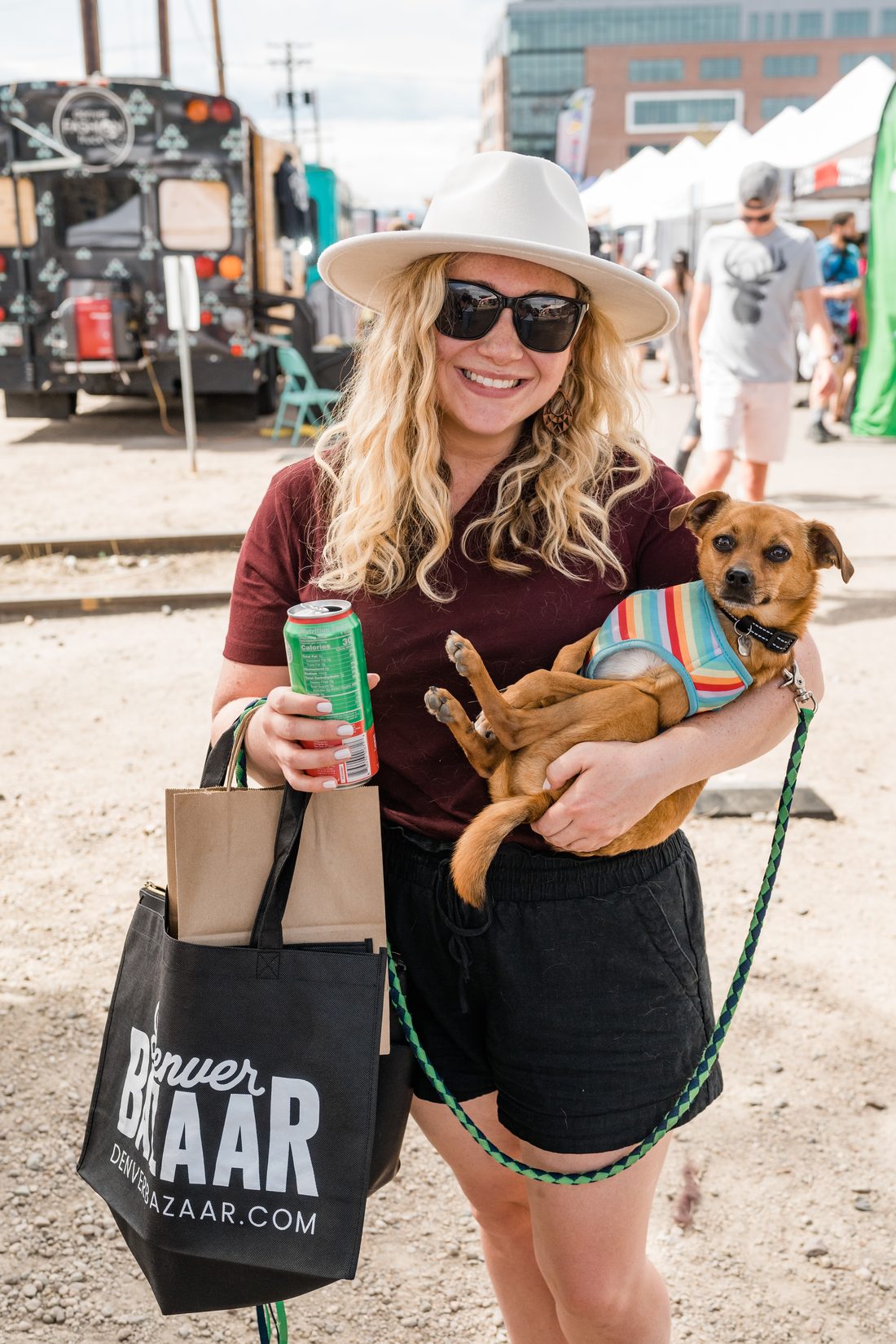 A shopper in a sun hat holds her small dog and a Denver BAZAAR tote.